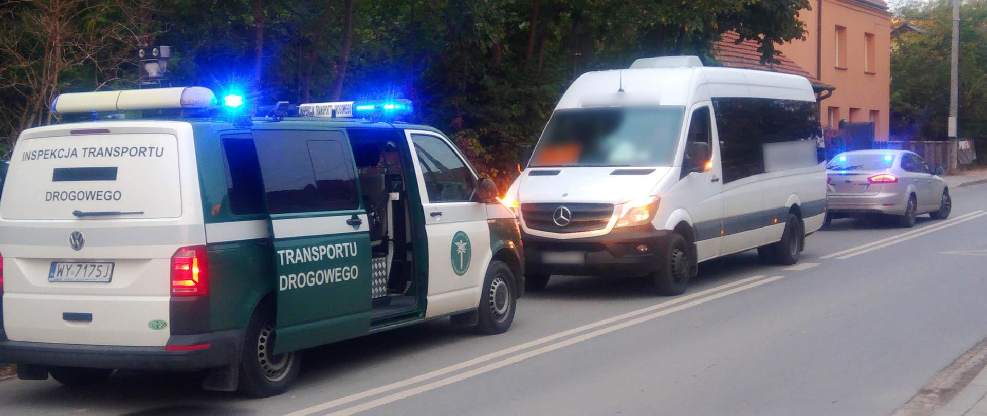 Przed biało-zielonym radiowozem typu bus z włączonymi niebieskimi sygnałami świetlnymi stoi biały autobus typu bus. Z tyłu nieoznakowany srebrny radiowóz z włączonymi niebieskimi światłami błyskowymi. 