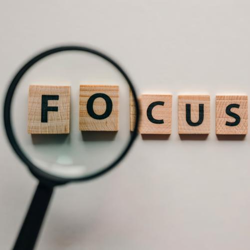 Na szarym tle leżą drewniane klocki z literami układające się w napis FOCUS . Pierwsze dwie litery powiększone przez lupę.