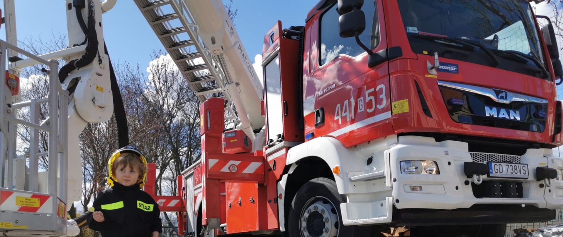 fotografia przedstawia chłopca w ubraniu strażaka, stojącego na placu komendy przy wozie strażackim