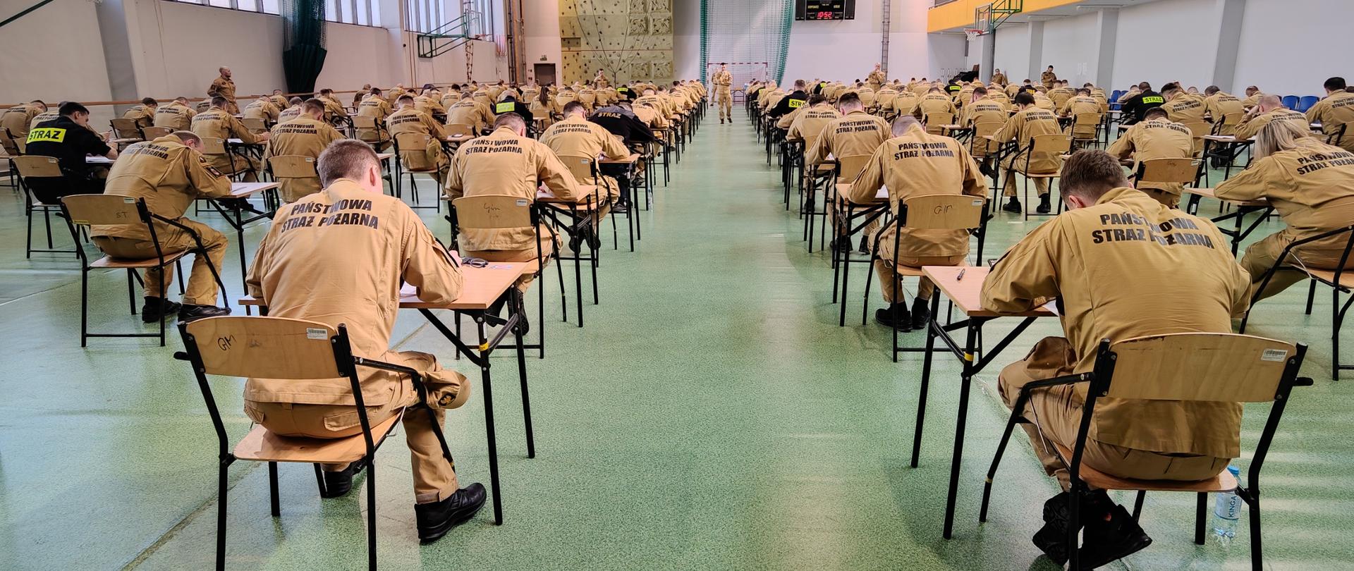 Wnętrze Sali gimnastycznej SA PSP w Krakowie, przy stolikach losujący numery miejsc strażacy w umundurowaniu służbowym (koloru musztardowego i w niebieskich koszulach). 