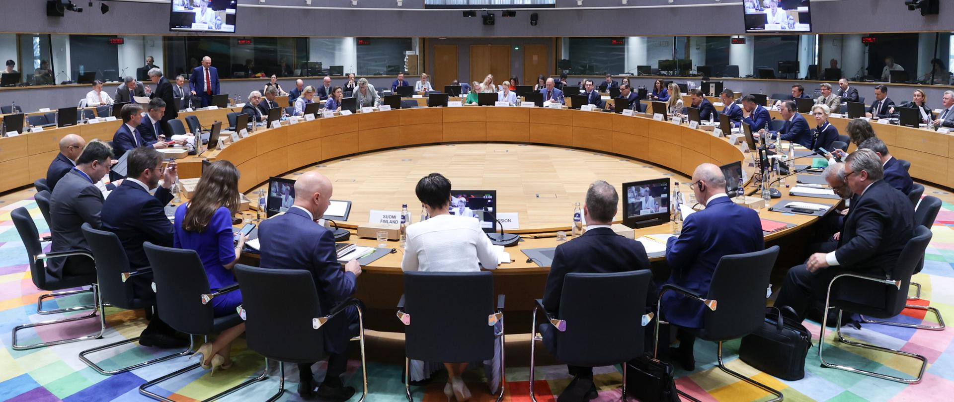 W budynku Europa w Radzie UE w nowoczesnej sali konferencyjnej uczestnicy spotkania siedzą przy dużym, okrągłym stole. Przed delegacjami znajdują się monitory, mikrofony, dokumenty oraz tabliczki z nazwami państw. W tle widoczne są kolejne stanowiska oraz kabiny tłumaczy. Charakterystyczny, wielobarwny sufit nadaje wnętrzu nowoczesny wygląd.