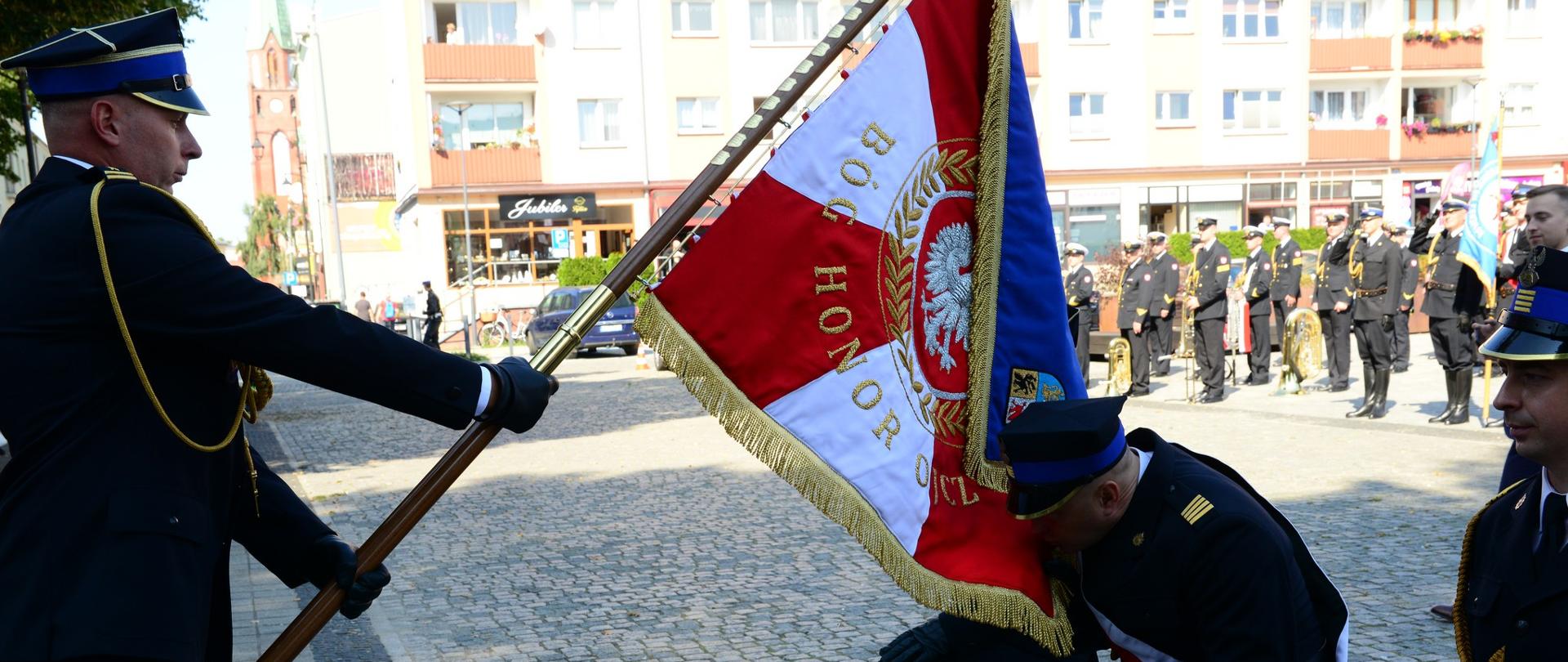 Komendant powiatowy Państwowej Straży Pożarnej w Lęborku pochyla sztandar, strażak z pocztu sztandarowego z Komendy Powiatowej Państwowej Straży Pożarnej w Lęborku klęczą, jeden ze funkcjonariuszy całuje sztandar. 