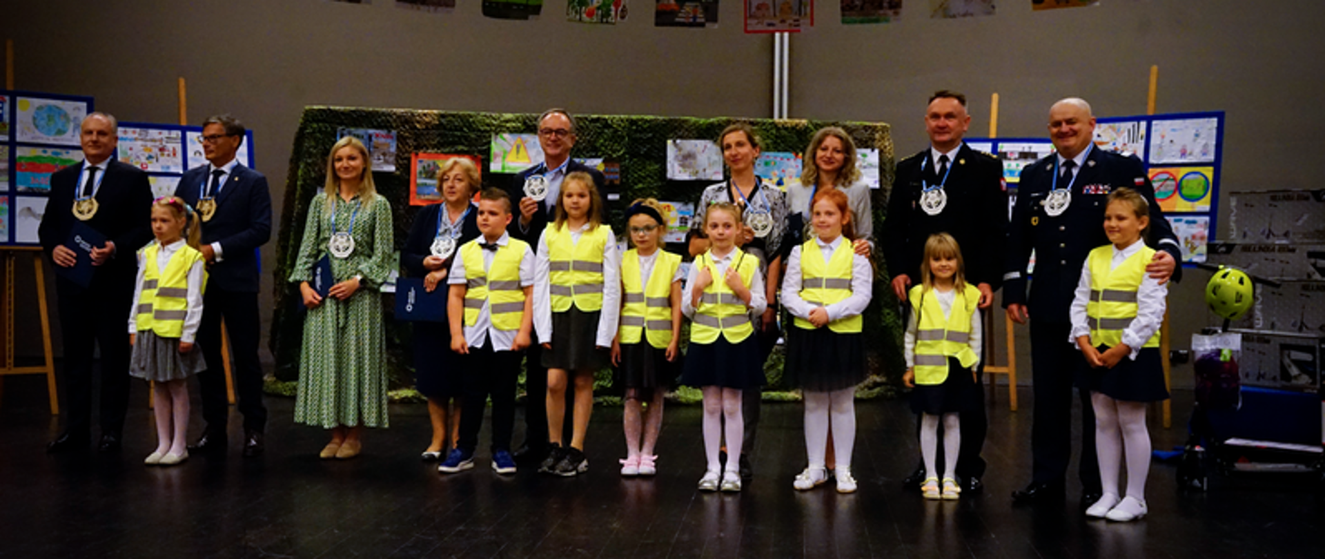 Dzieci ubrane w kamizelki odblaskowe stoją wspólnie z wojewodą pomorskim, przedstawicielami Policji, Państwowej Straży Pożarnej oraz sponsorami. Za nimi znajdują się tablice z rysunkami.