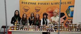 Sześć kobiet stoi za drewnianym ladą stoiska „Rodzinne Pasieki Warcholak”. Na blacie ustawione są dziesiątki słoików z miodem o różnych odcieniach, a w tle widać baner z motywem pszczół.