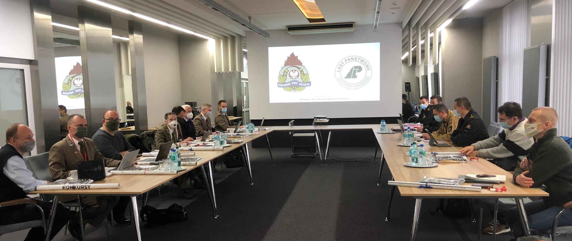 Uczestnicy spotkania w sprawie wymagań dotyczących ochrony przeciwpożarowej lasów przy stole ułożonym "w podkowę" W tle ekran z prezentacją