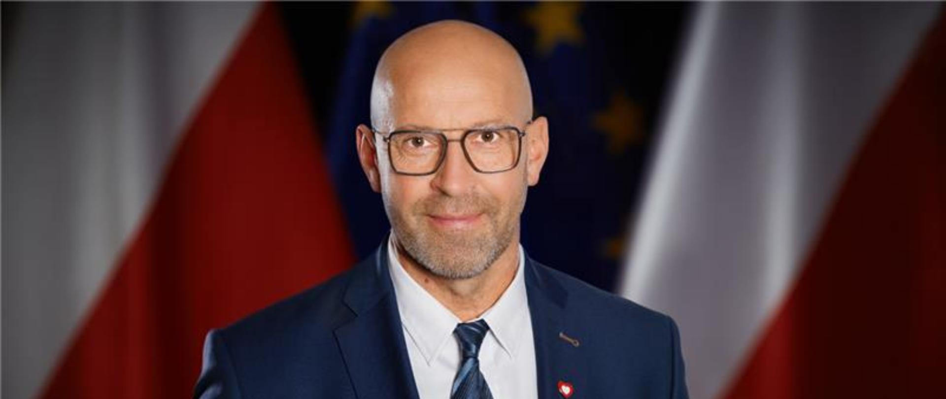 Minister Sportu i Turystyki Jakub Rutnicki stoi na tle flag Polski i Unii Europejskiej