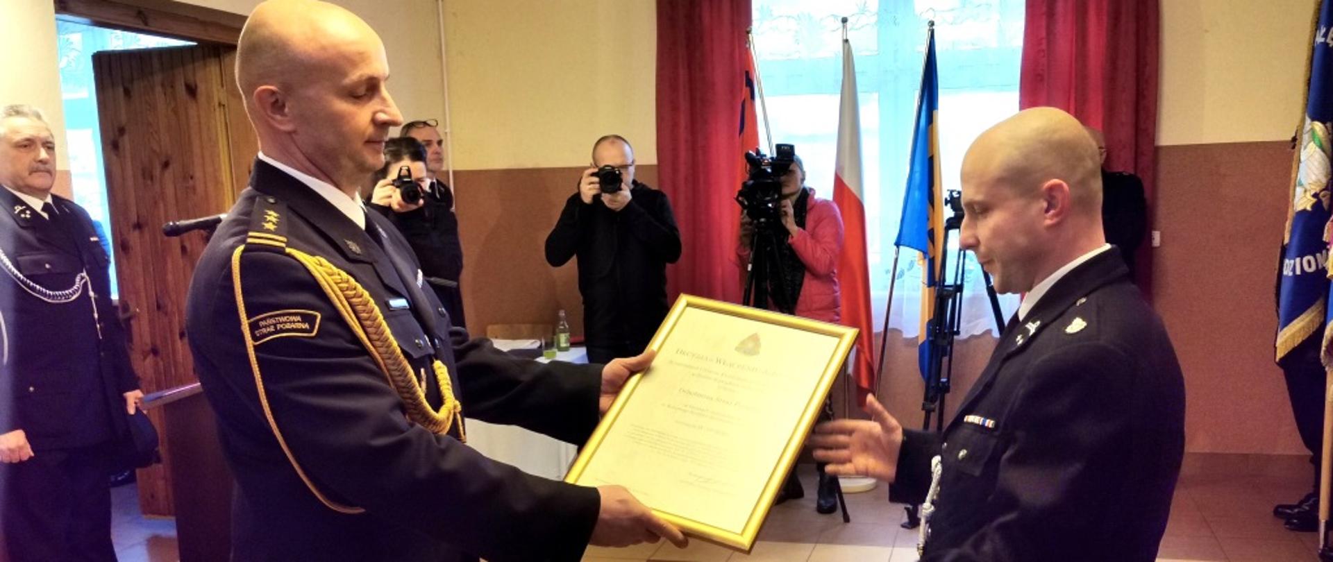 Zdjęcie przedstawia osobę Komendanta Miejskiego Państwowej Straży Pożarnej w Krośnie u mundurze galowym, wręczającego decyzję o włączeniu jednostki OSP Łączki Jagiellońskie do Krajowego Systemy Ratowniczo-Gaśniczego. Decyzję odbiera stojący naprzeciwko prezes OSP Łączki Jagiellońskie, występujący w mundurze galowym.