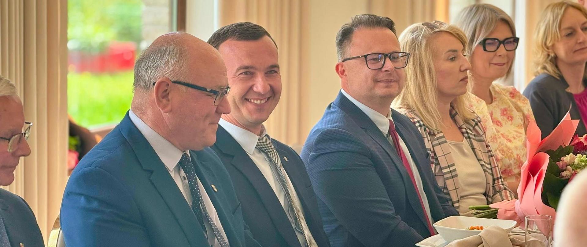 Kilkanaście osób siedzi przy stole. Na stole znajdują się naczynia i jedzenie. 