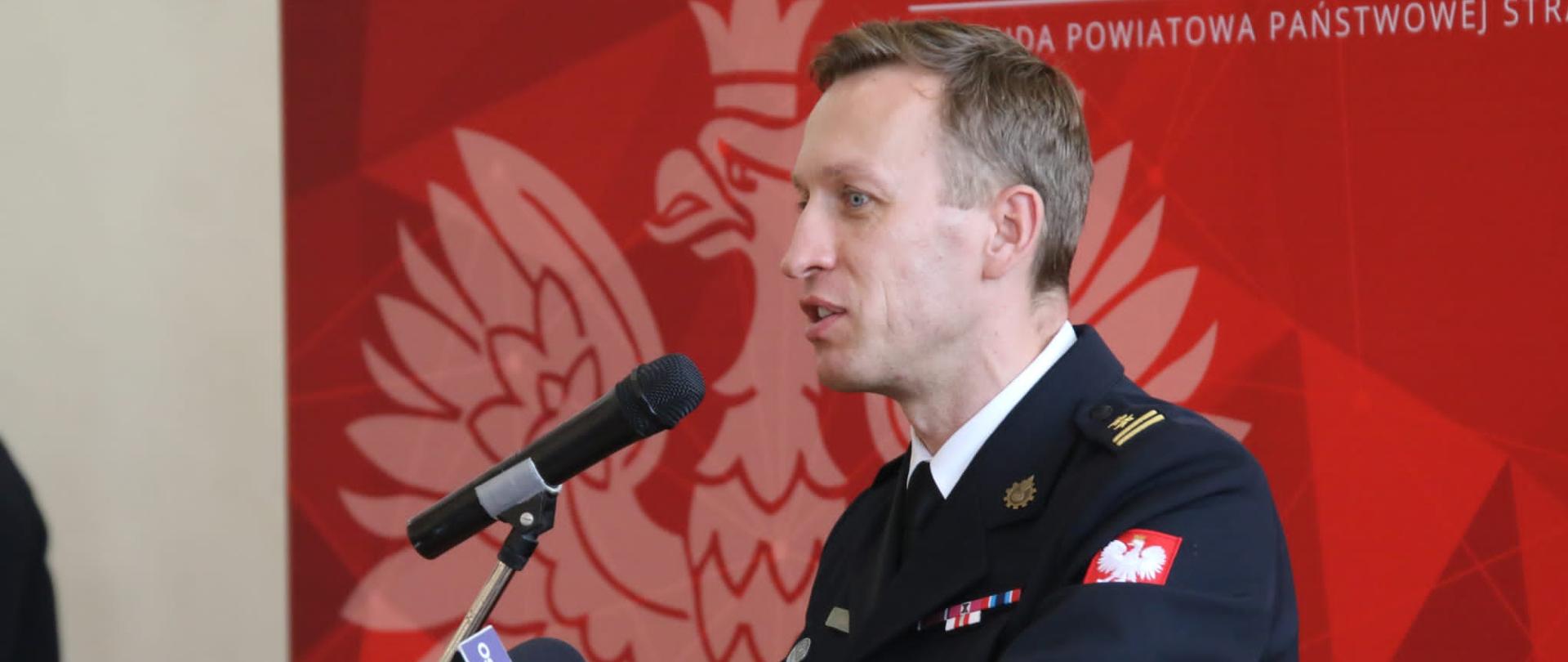 strażak stojacy przy mównicy, za nim czerwone tło