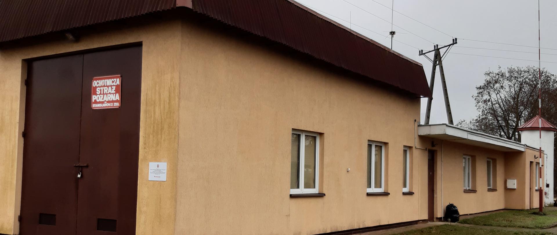 Strażnica OSP Stanisławowo Zegrzyńskie. Na zdjęciu budynek murowany kryty blachą parterowy. Dach i brama garażowa brązowa z prawej strony maszt flagowy.