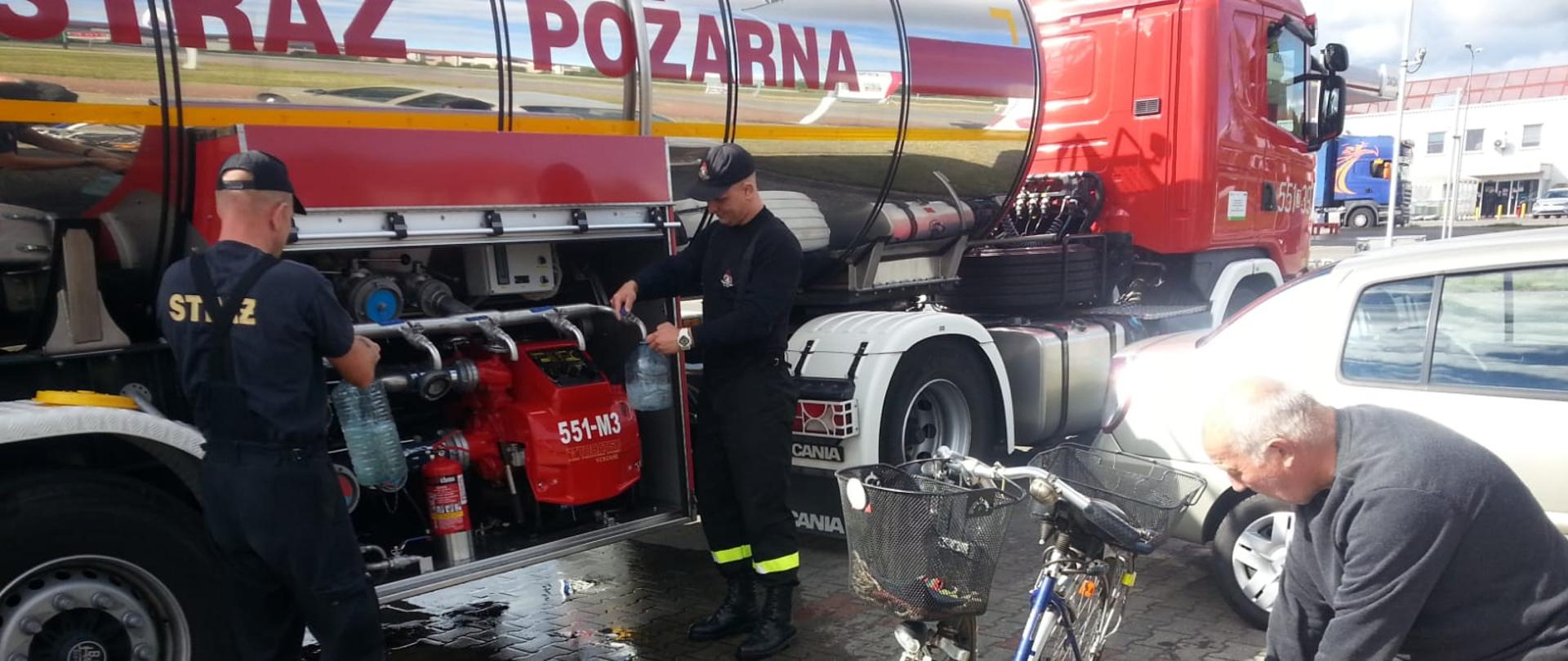 Zdjęcie przedstawia cysternę strażacką wraz ze strażakami którzy napelniają pojemniki mieszkańcom wodą.