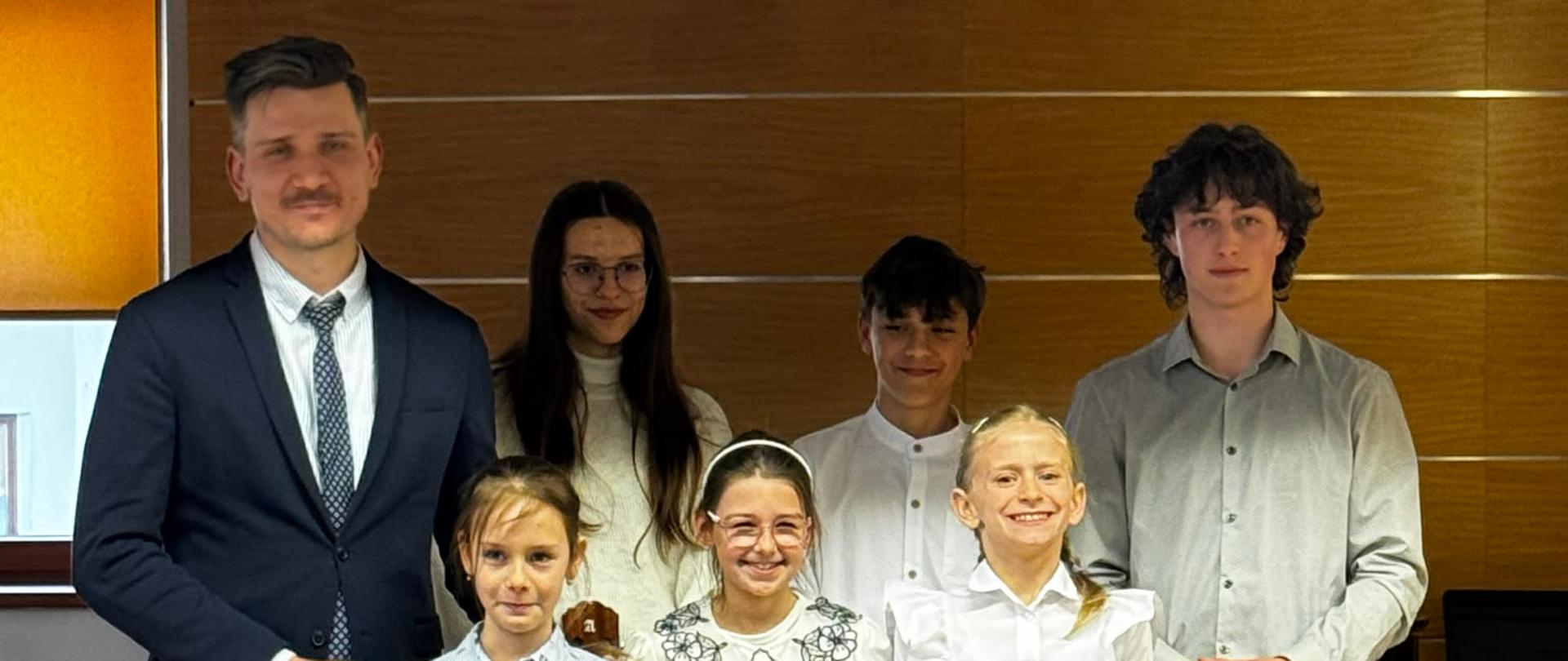 Zdjęcie przedstawia grupę siedmiu osób – prawdopodobnie uczniów i ich nauczyciela – pozujących po występie lub lekcji gry na instrumencie. Postacie: Na pierwszym planie stoją trzy uśmiechnięte dziewczynki, a obok nich (po lewej) wysoki mężczyzna w garniturze, który trzyma dłoń na ramieniu jednej z nich. W drugim rzędzie widać dwoje nastolatków oraz młodą kobietę. Instrumenty: Czworo uczniów trzyma przed sobą klasyczne gitary akustyczne. Instrumenty różnią się nieco rozmiarem, co sugeruje, że są dopasowane do wzrostu młodych muzyków. Ubiór: Wszyscy są ubrani w stroje galowe. Dominują białe koszule, bluzki z ozdobnymi kołnierzykami oraz ciemne spodnie. Mężczyzna ma na sobie ciemnogranatową marynarkę i krawat. Tło: Grupa znajduje się w jasnym pomieszczeniu, prawdopodobnie w szkole muzycznej lub sali koncertowej. Ściana za nimi jest wyłożona drewnianymi panelami, co jest typowe dla sal o dobrej akustyce. To bardzo sympatyczna fotografia, która emanuje dumą i pasją do muzyki.