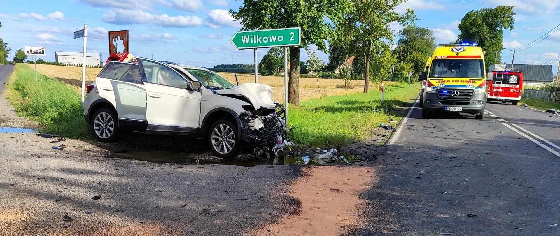 Uszkodzony biały samochód na poboczu, po prawej karetka pogotowia na drodze