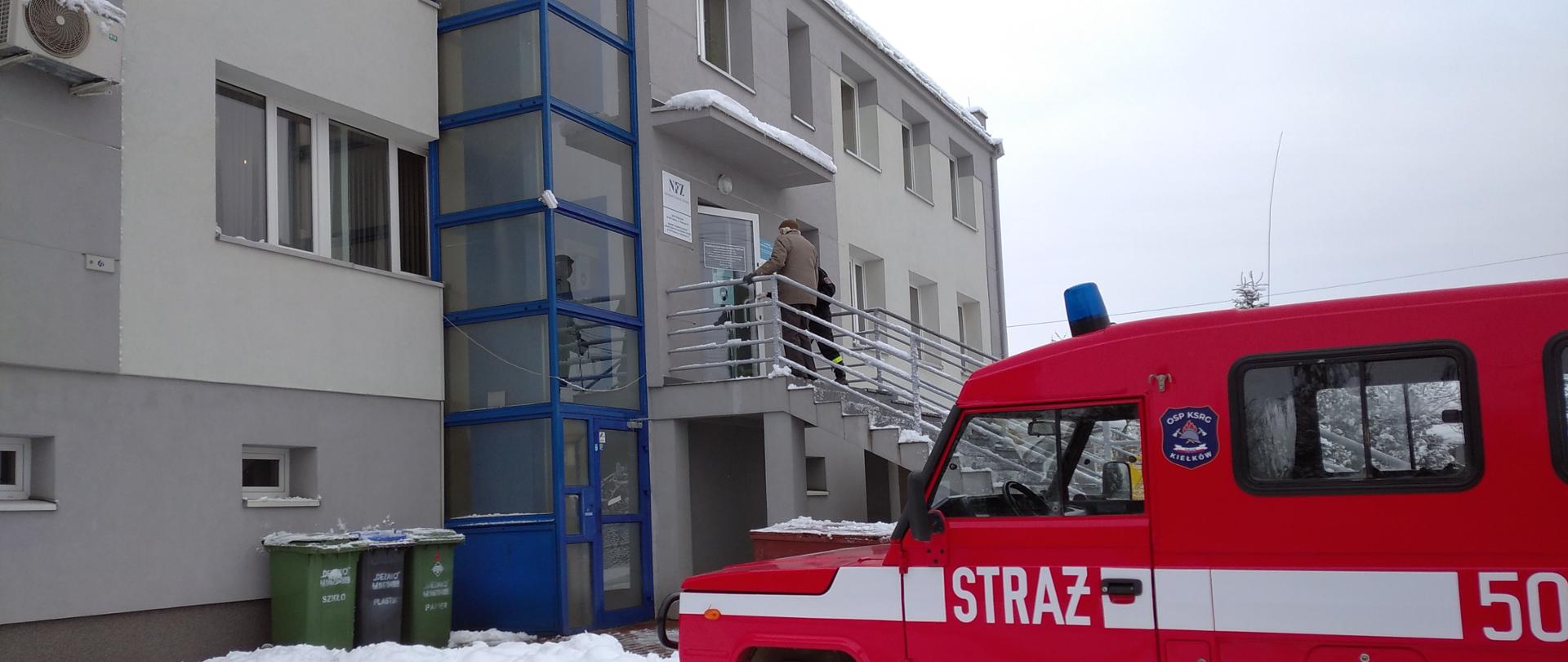 Na zdjęciu widać terenowy, osobowy samochód strażacki i osobę w podeszłym wieku wchodzącą ze strażakiem do ośrodka zdrowia