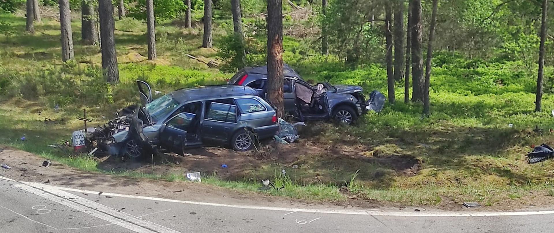 Na zdjęciu na poboczu drogi przy drzewie znajdują się dwa uszkodzone pojazdy osobowe koloru szarego z widocznymi uszkodzeniami.