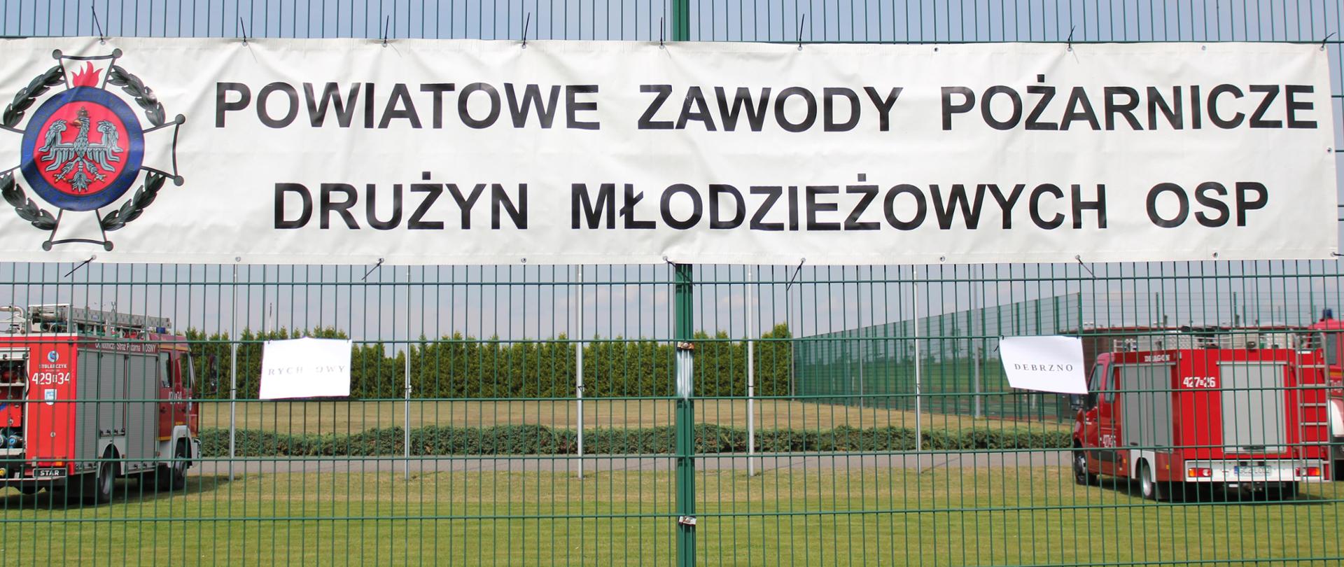 Zdjęcie przedstawia baner: " POWIATOWE ZAWODY POŻARNICZE DRUŻYN MŁODZIEŻOWYCH OSP". W tle samochody pożarnicze.