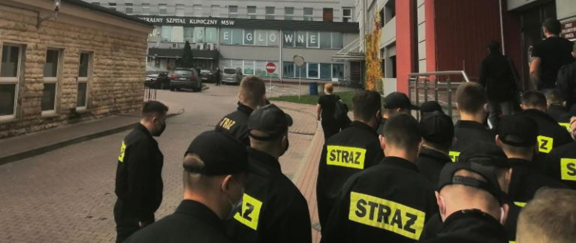 Grupa strażaków, stojąca tyłem w ubraniach koszarowych na placu przed Centralnym Szpitalem Klinicznym MSWiA w Warszawie.