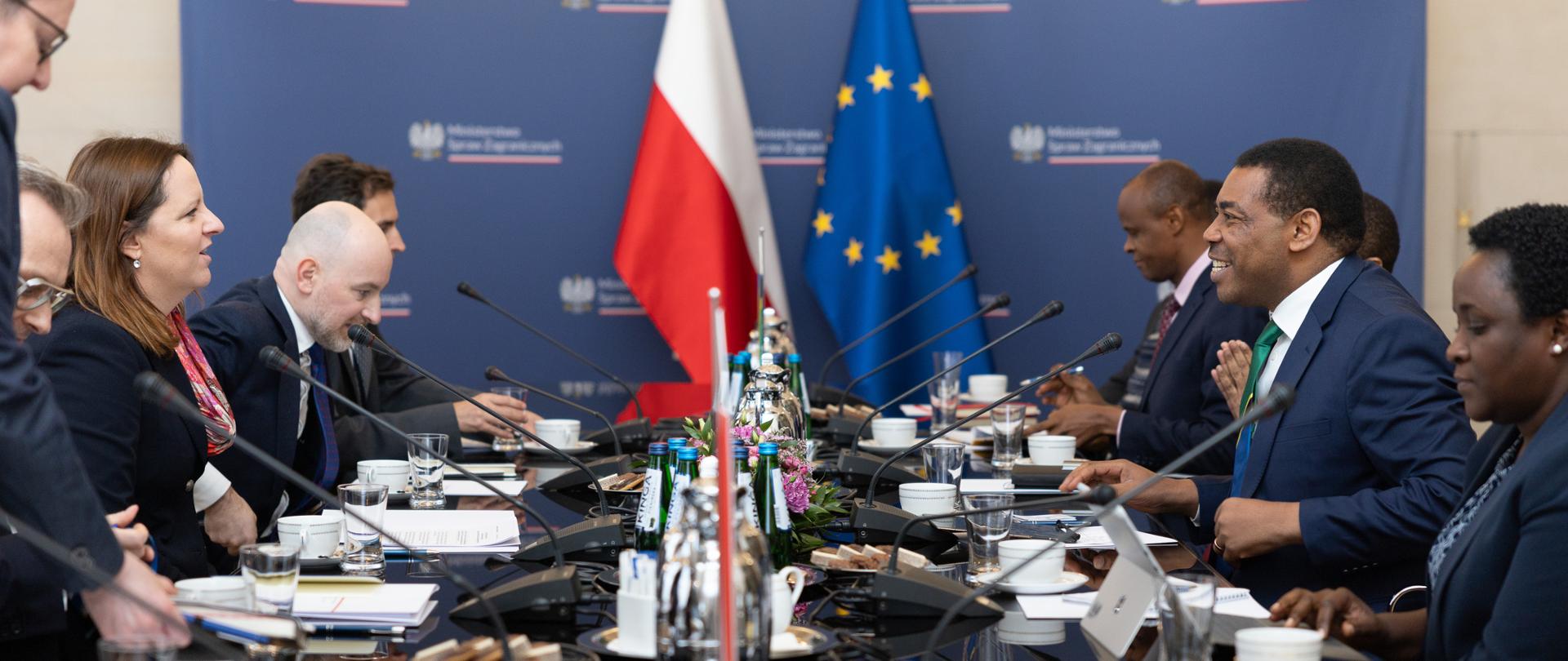 Na zdjęciu jest ok. 10 osób, które siedzą przy stole, na którym są mikrofony, zastawa stołowa i zeszyty. W tle są flagi Polski i Unii Europejskiej. 