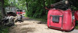 Zdjęcie przedstawia miejsce wypadku na drodze wojewódzkiej nr 297. Na zdjęciu widoczny jest samochód ciężarowy po wypadku - oderwana kabina pojazdu leży na drodze.W tle widoczny jest samochód z Jednostki Ratowniczo-Gaśniczej z Lwówka Śląskiego. 