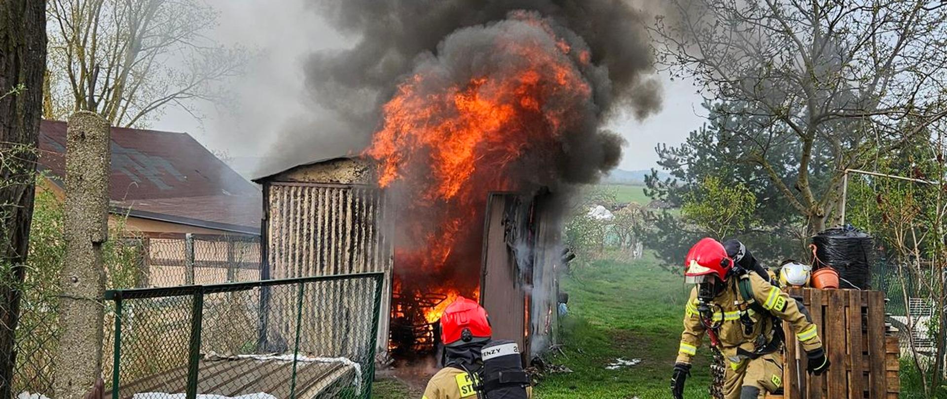 Zdjęcie przedstawia strażaków w trakcie gaszenia pożaru garażu.
W tle samochód strażacki, budynek, drzewa.
