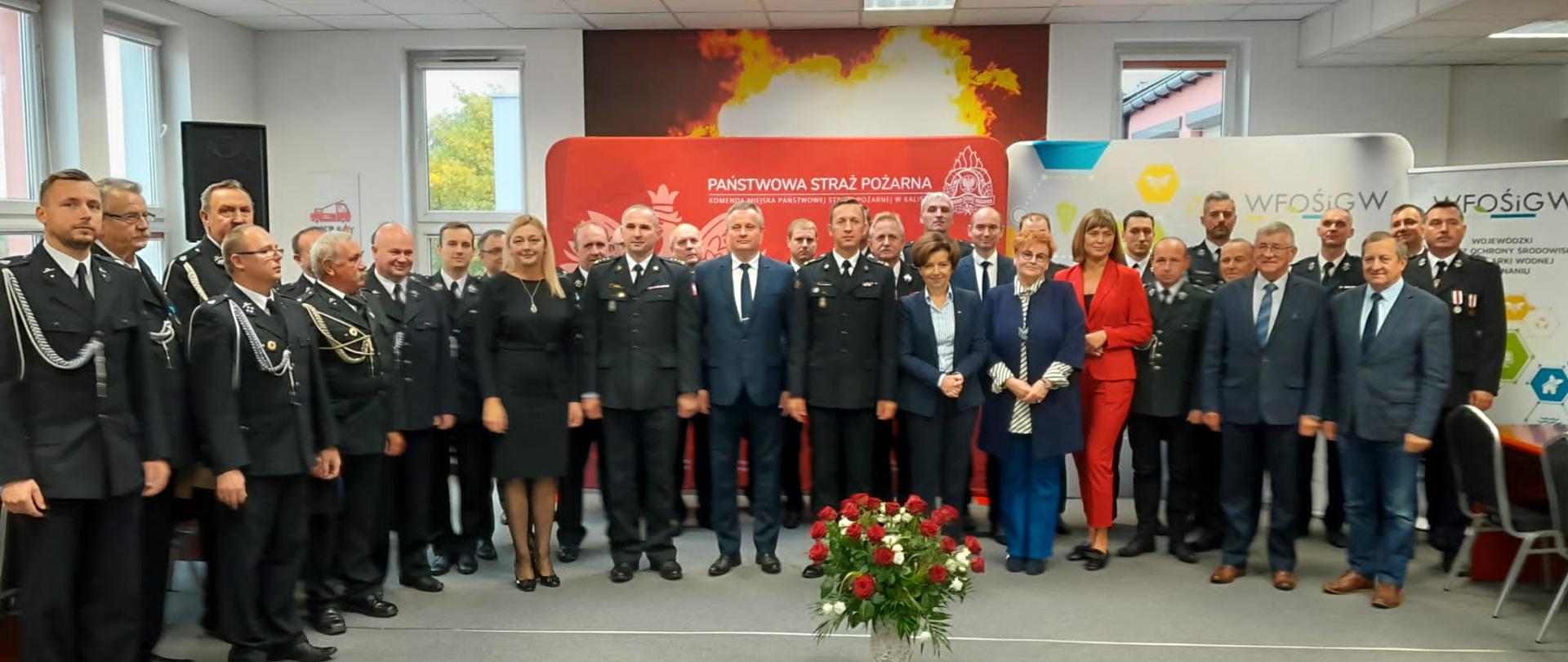 Zdjęcie wykonane w pomieszczeniu Komendy Miejskiej Państwowej Straży Pożarnej w Kaliszu. Na zdjęciu widać druhów OSP w umundurowaniu wyjściowym, funkcjonariuszy PSP w umundurowaniu wyjściowym oraz zaproszonych gości. Pomieszczenie sztucznie oświetlone. Na podłodze szary dywan. Jasne ściany. Przy ścianach banery Państwowej Straży Pożarnej oraz Wojewódzkiego Funduszu Ochrony środowiska i Gospodarki Wodnej.