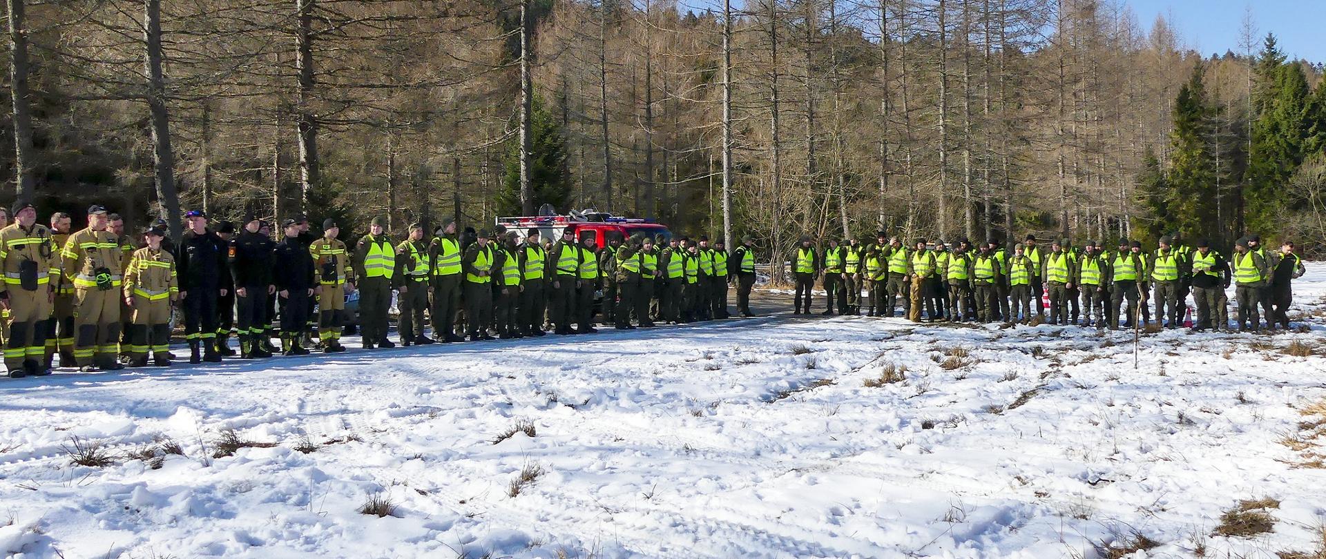 Zdjęcie przedstawia strażaków OSP oraz żołnierzy którzy ustawieni są w szyku. Drugi szyk stoi równolegle do pierwszego. Za żołnierzami widać część samochodu strażackiego. 
