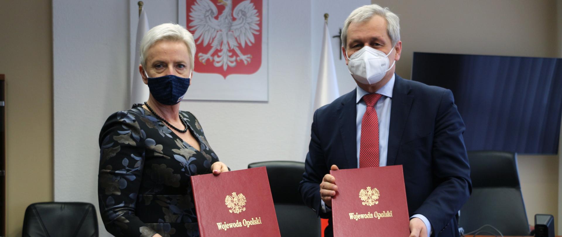 Na zdjęciu od prawej strony wojewoda opolski, obok prezydent Kędzierzyna-Koźla. Stoją obok siebie, pozują do aparatu, w dłoniach każde z nich trzyma teczkę bordową z napisem wojewoda opolski oraz złotym orzełkiem. Za nimi widać biurko w gabinecie wojewody, wysokie czarne fotele. Na ścianie za ich plecami godło, oraz biało-czerwona flaga. Po prawej stronie kadru - w tle, widać duży monitor telewizora. 