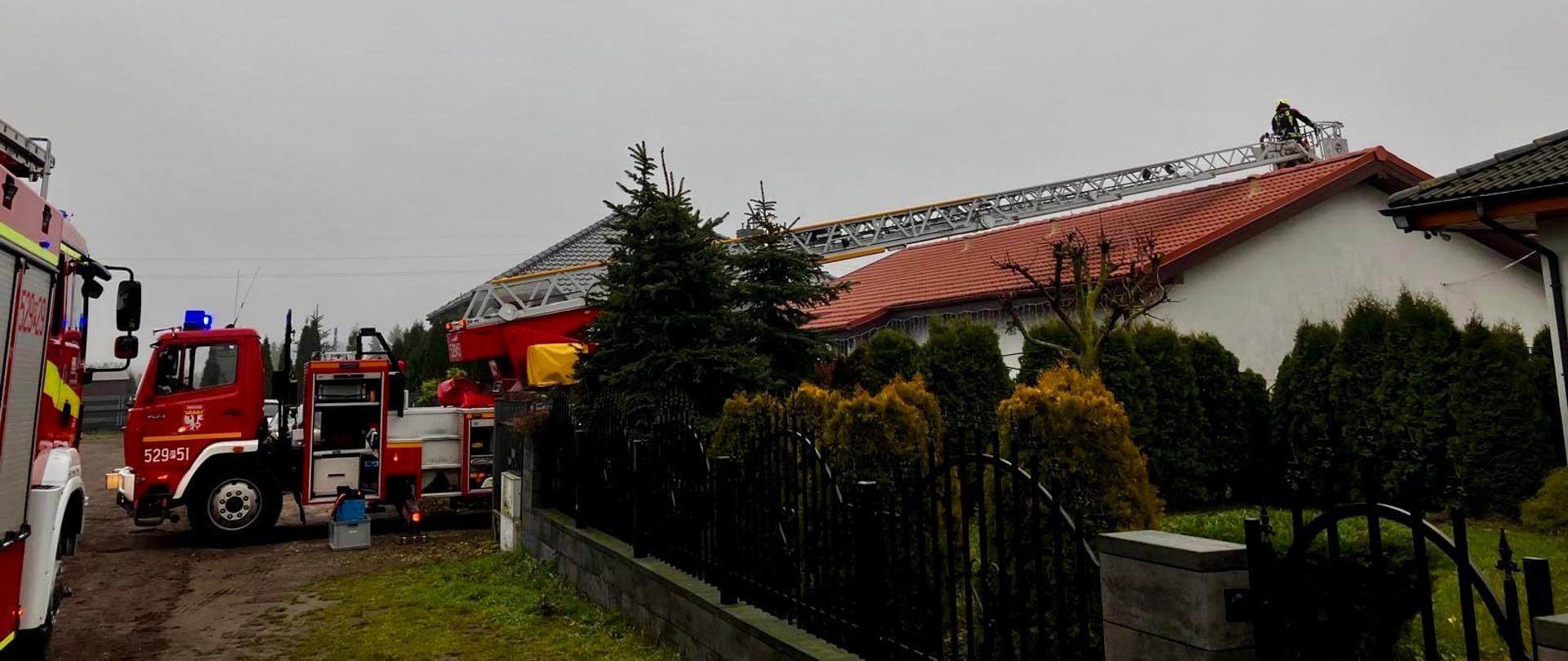 Akcja straży pożarnej przy domu jednorodzinnym. Wóz strażacki z wysuniętą drabiną mechaniczną sięga dachu budynku, na którym znajduje się strażak. Wokół posesji widać ogrodzenie, drzewa i ogród, a pogoda jest pochmurna