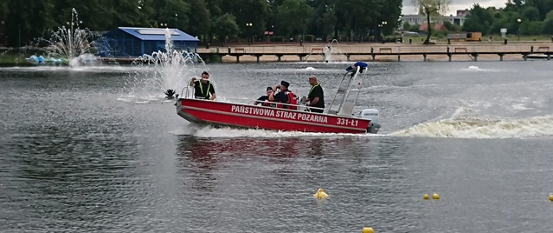 Zdjęcie wykonane na terenie zbiornika wodnego „Borki” w Radomiu. Na zdjęciu widać łódź ratowniczą należąca do Komendy Miejskiej Państwowej Straży Pożarnej w Radomiu. Łódź w kolorze czerwono – srebrnym. Na boku posiada biały napis Państwowa Straż Pożarna. W łodzi znajduje się mieszany patrol złożony z Policjantów i funkcjonariuszy Państwowej Straży Pożarnej w Radomiu. Jeden z Policjantów w rękach trzyma lornetkę wypatrując niebezpiecznych zachowań. 