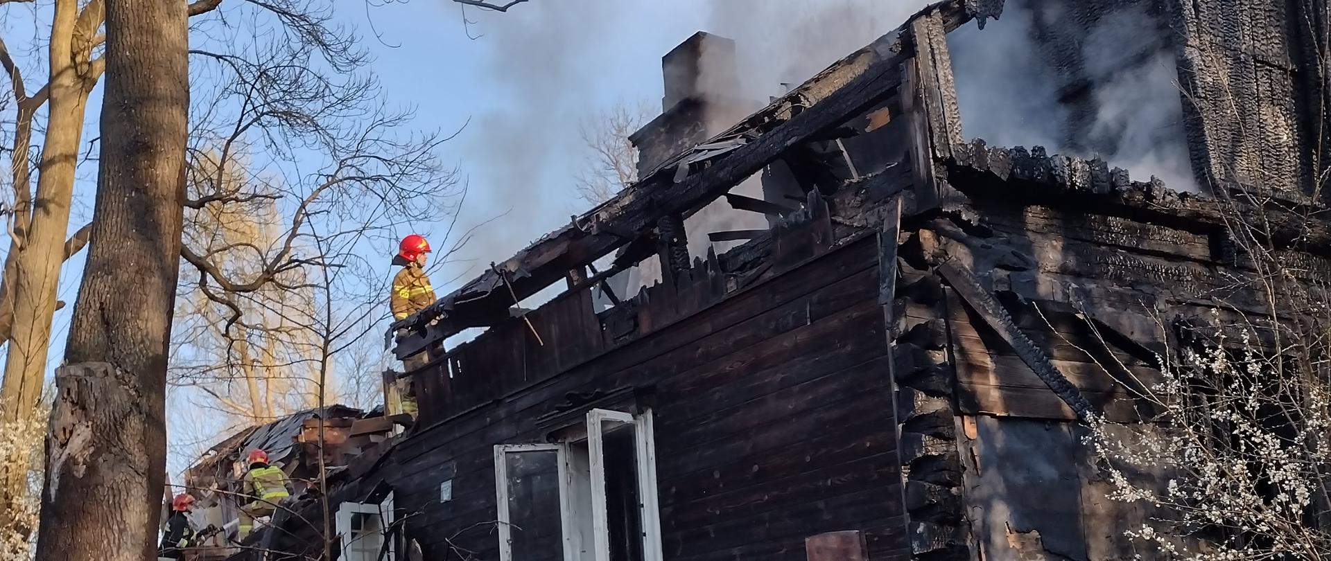Spalony drewniany pustostan po budynku mieszkalnym. Na dachu pracujący strażak.