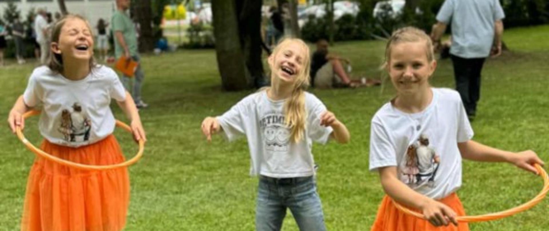Zdjęcie kolorowe. Trzy dziewczynki bawią się hula-hop w parku. Dwie z nich mają pomarańczowe spódnice i białe koszulki. Trzecia dziewczynka ma jeansy i białą koszulkę. Wszystkie uśmiechają się. W tle widać drzewa i inne osoby.