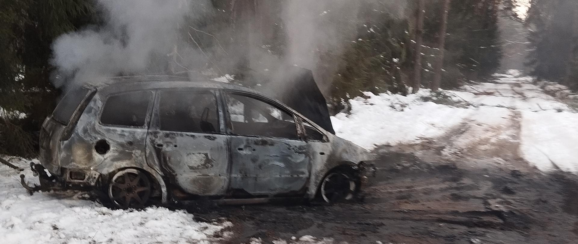 Spalony samochód stoi na drodze leśnej. Na drodze i poboczu widać śnieg. Z samochodu unosi się dym