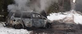 Spalony samochód stoi na drodze leśnej. Na drodze i poboczu widać śnieg. Z samochodu unosi się dym