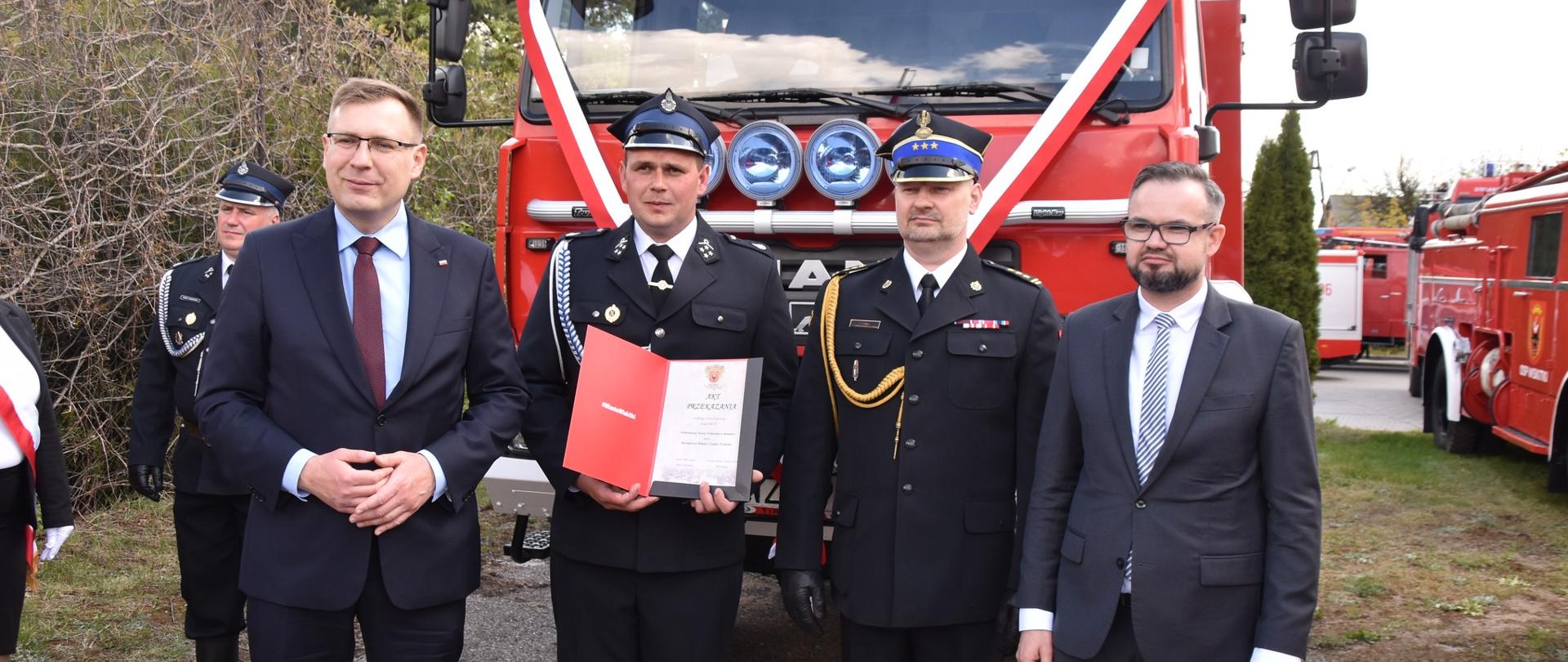 Komendant powiatowy PSP w Żyrardowie st. bryg. Tomasz Cybul stoi w towarzystwie burmistrza miasta i gminy Wiskitki Rafała Mitury, wiceministra Aktywów Państwowych Macieja Małeckiego oraz druha z OSP Jesionka. Ten ostatni trzyma z rękach akt przekazania samochodu pożarniczego. W tle samochód pożarniczy.