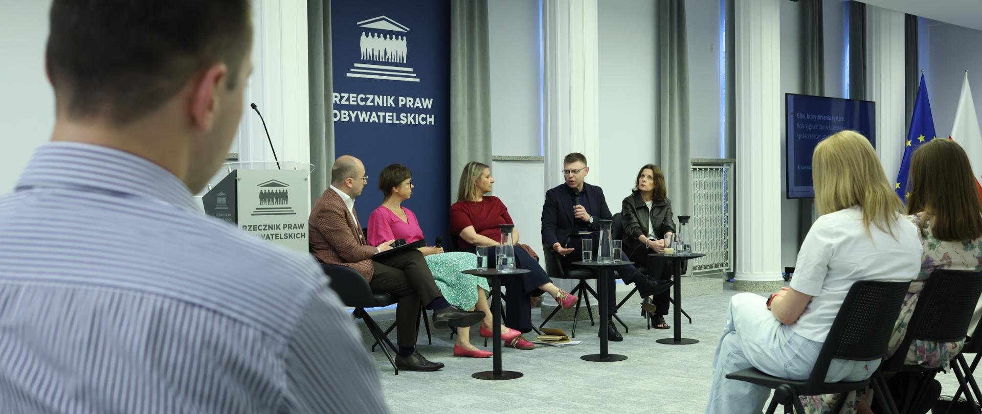 Pięć osób siedzi przy stolikach w sali konferencyjnej, jedna z nich mówi do mikrofonu. Rozmowie przysłuchują się osoby siedzące na miejscach dla publiczności