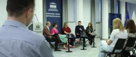 Pięć osób siedzi przy stolikach w sali konferencyjnej, jedna z nich mówi do mikrofonu. Rozmowie przysłuchują się osoby siedzące na miejscach dla publiczności