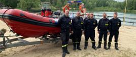 Stoją na piaszczystej plaży nad zbiornikiem wodnym. Za nimi znajduje się duża, czerwono-czarna łódź ratownicza typu RIB na przyczepie transportowej. W tle widać las oraz pomost ciągnący się wzdłuż brzegu. Niebo jest zachmurzone.