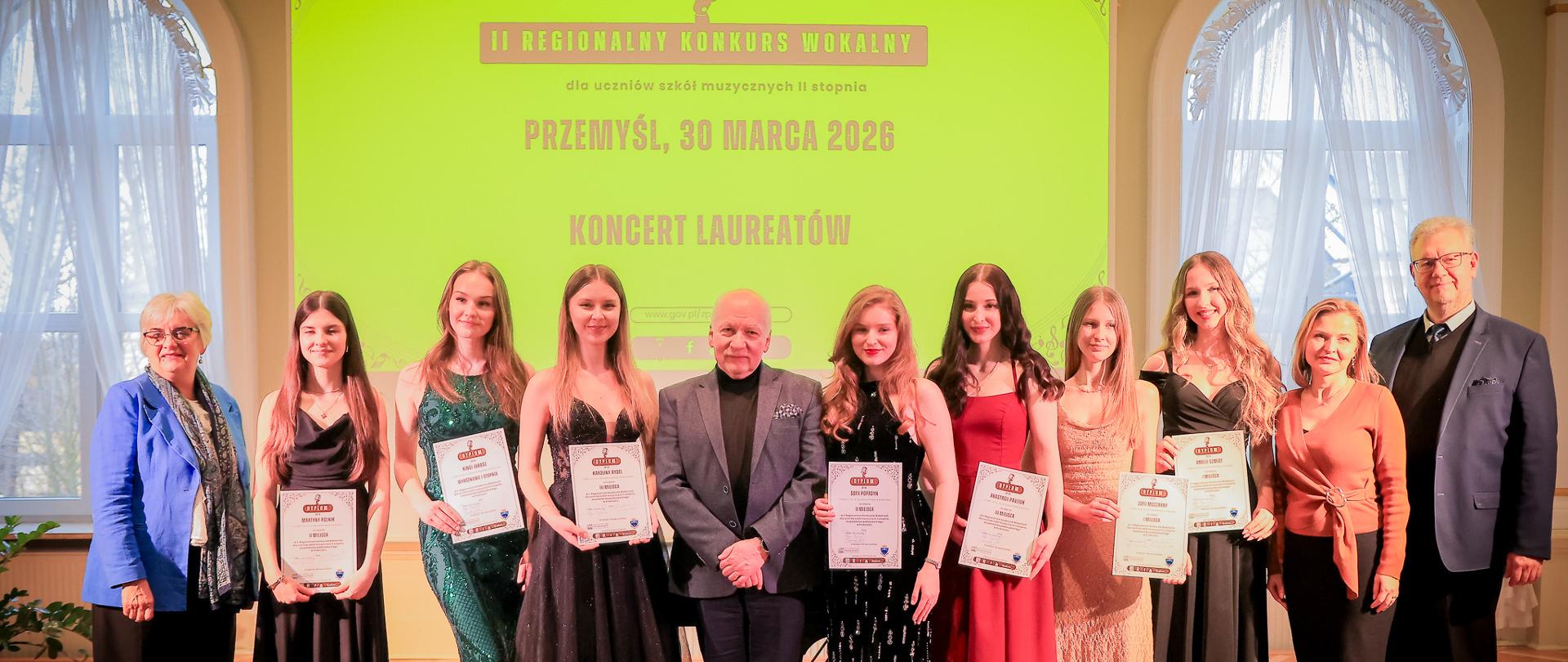 Grupa kilkunastu osób stoi na scenie podczas uroczystości w sali koncertowej. W centrum znajduje się starszy mężczyzna w garniturze, a po jego obu stronach stoją młode kobiety w eleganckich sukniach wieczorowych, trzymające dyplomy. Po bokach stoją również dorośli opiekunowie lub organizatorzy. W tle widoczny jest duży ekran z napisem o „II Regionalnym Konkursie Wokalnym” oraz datą i miejscem wydarzenia