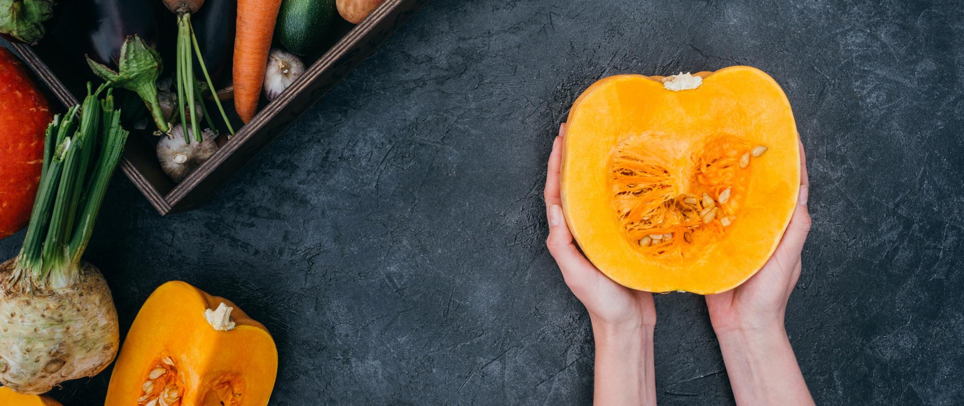 Na zdjęciu w około są różne warzywa. W centrum jest przepołowiona dynia trzymana w dłoniach.