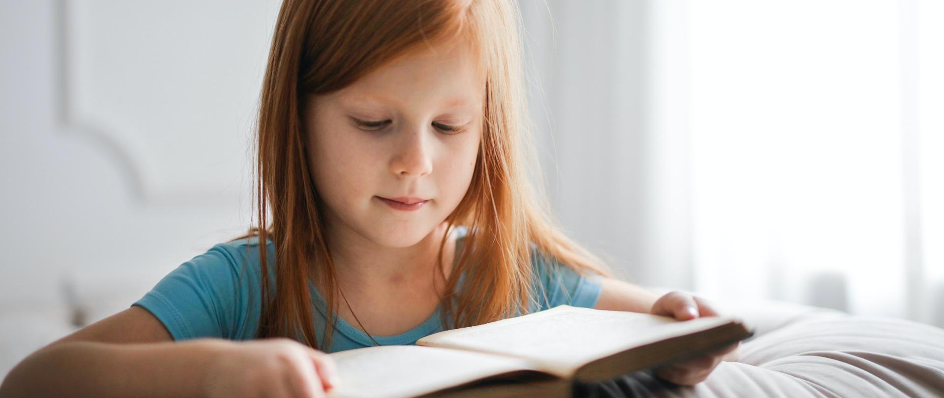 Zdjęcie w poziomie. Ruda dziewczynka w niebieskiej bluzce czytająca książkę.
