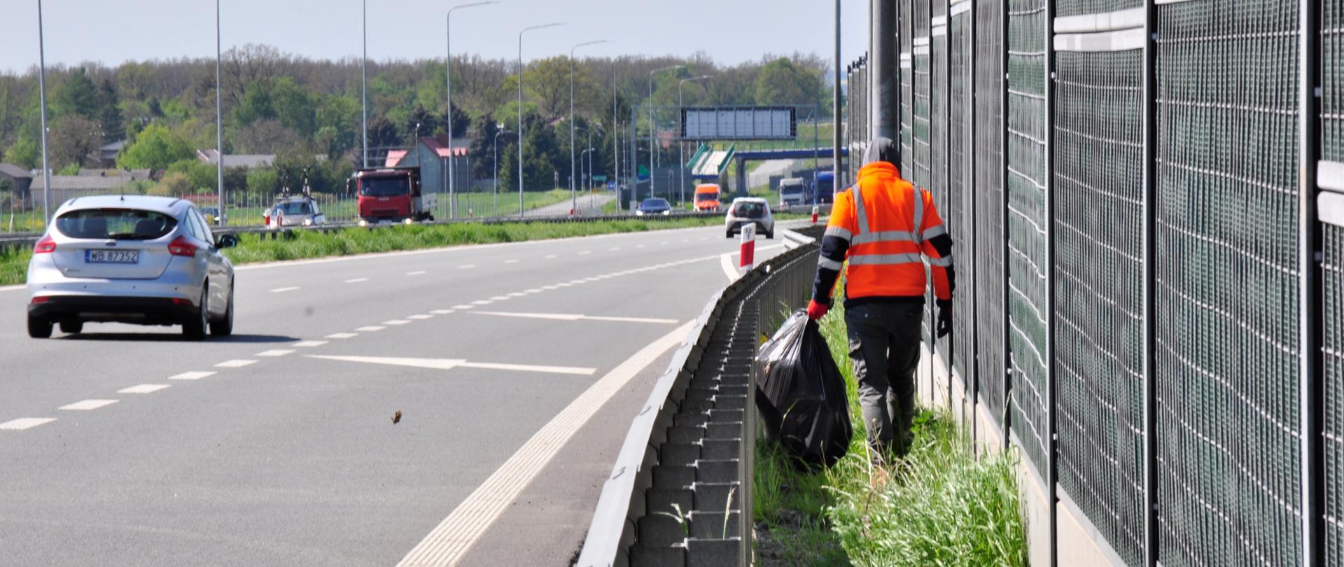 Zdjęcie przedstawia osobę ubraną w pomarańczową kamizelkę odblaskową, która porusza się wzdłuż pobocza autostrady, zbierając śmieci. Osoba ta trzyma duży czarny worek na śmieci i idzie obok metalowego ogrodzenia dźwiękochłonnego. Na drodze w tle widoczne są pojazdy, w tym srebrny samochód osobowy oraz czerwona ciężarówka. Po lewej stronie zdjęcia znajduje się pas zieleni oddzielający jezdnię od pobocza. W tle widoczne są drzewa oraz budynki, a dzień jest pogodny, z lekkim zachmurzeniem.