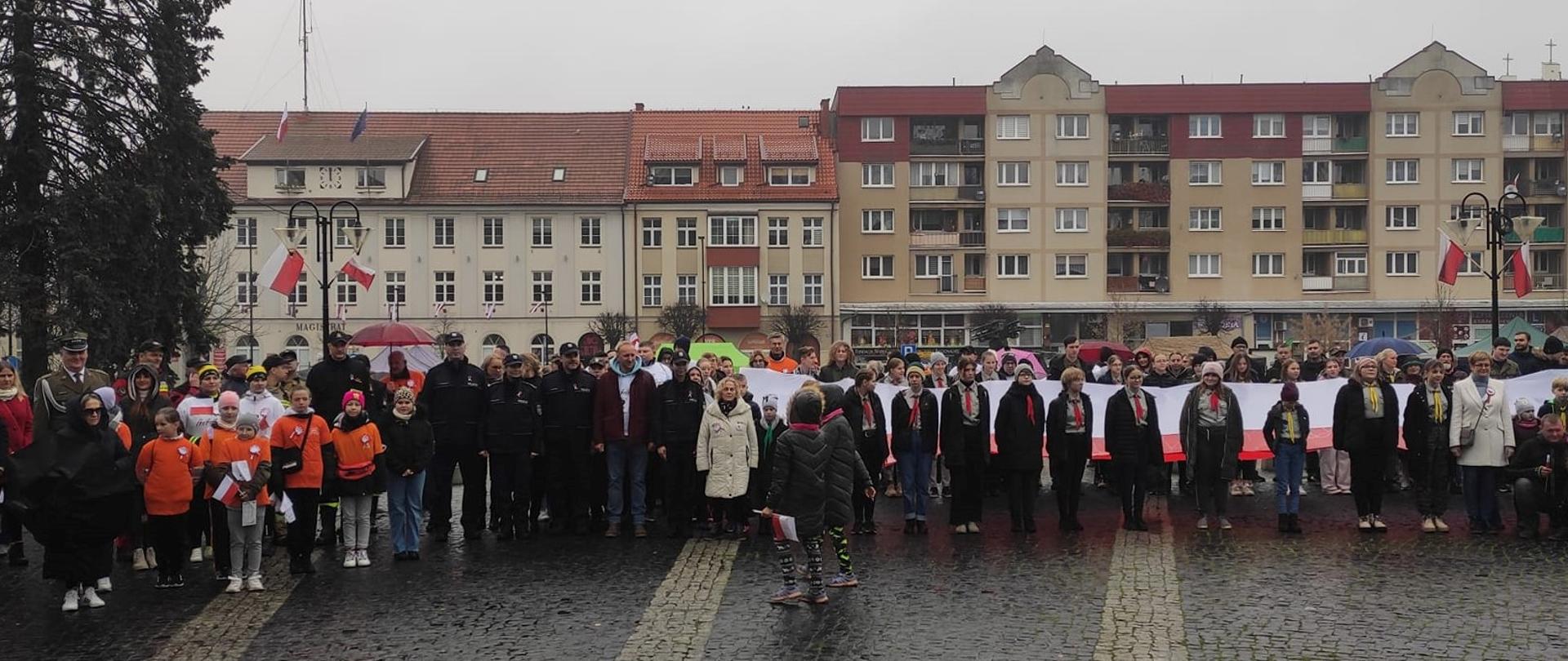 Zdjęcie przedstawia funkcjonariuszy Komendy Powiatowej Państwowej Straży Pożarnej w Gołdapi, którzy o godz. 12:00 na Placu Zwycięstwa w Gołdapi (nieopodal Pomnika Niepodległości) wspólnie z mieszkańcami miasta oraz przedstawicielami władz samorządowych i innych służb zaśpiewali hymn państwowy w ramach akcji „Niepodległa do Hymnu”. Uczestnicy uroczystości trzymają flagę Rzeczypospolitej Polskiej
