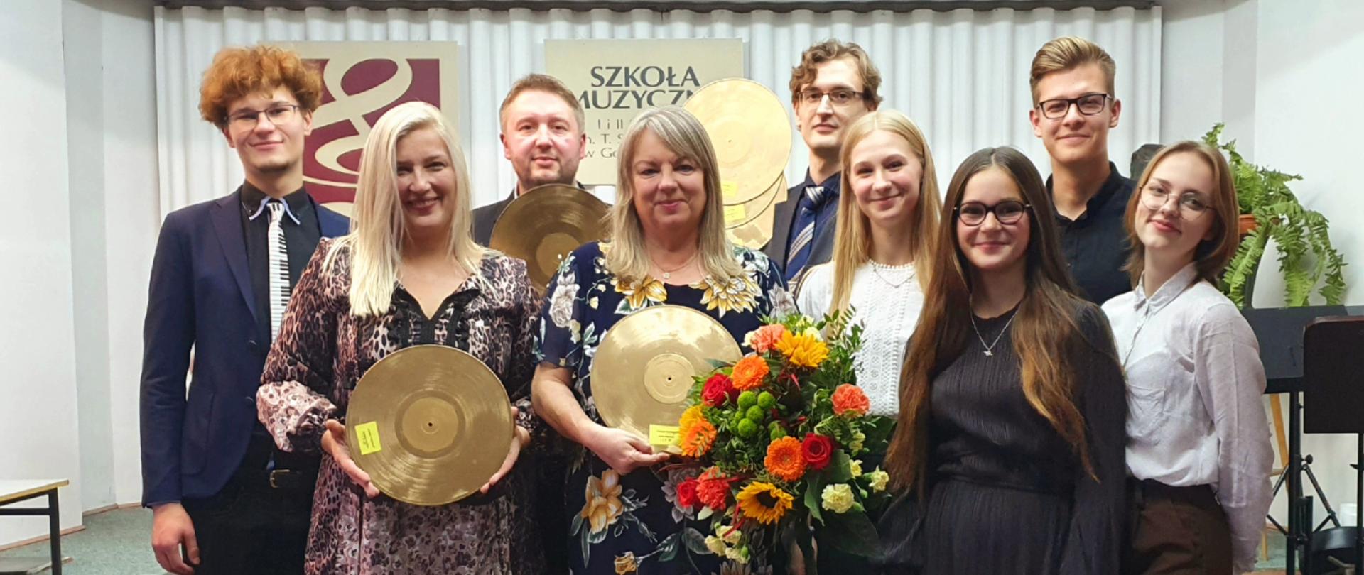 Zdjęcie nauczycieli i uczniów w szkolnej auli. Nauczyciele trzymają złote płyty, tzw. Tadeusze przyznawane przez uczniów
