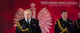 To zdjęcie przedstawia oficjalną uroczystość strażacką, utrzymaną w podniosłym tonie. Głównym punktem kompozycji jest przemawiający funkcjonariusz. Mężczyzna w ciemnym mundurze galowym Państwowej Straży Pożarnej (odchodzący na zaopatrzenie emerytalne Komendant Powiatowy PSP w Wołowie stoi przy jasnej, drewnianej mównicy. Na prawym ramieniu ma gruby, złoty sznur galowy, a na piersi widoczne są baretki odznaczeń oraz plakietka imienna. Przemawia do mikrofonu zamontowanego na statywie, trzymając w dłoniach kartkę z tekstem. Na przedniej części mównicy widnieje kolorowe logo straży pożarnej. Za plecami mówcy znajduje się duża, bordowa ścianka o geometrycznym wzorze. Umieszczono na niej: Duży, biały kontur Godła Polski. Biały napis: „Państwowa Straż Pożarna Komenda Powiatowa PSP w Wołowie”. Po prawej stronie zdjęcia widać Dolnośląskiego Komendanta Wojewódzkiego PSP w identycznym mundurze galowym, który stoi nieruchomo w postawie zasadniczej, przysłuchując się wystąpieniu.