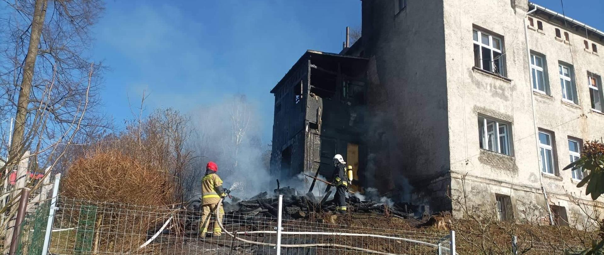 Na fotografii widać budynek po pożarze. Budynek mieszkalny koloru szarego. Pożar przybudówki przy budynku. Zdjęcie wykonane w dzień. Na zdjęciu znajdują się także strażacy. 