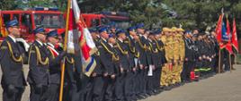 Panoramiczne zdjęcie przedstawiające długi szereg strażaków stojących na baczność podczas oficjalnego apelu. Szereg ciągnie się ukośnie od lewego dolnego rogu w głąb prawego boku zdjęcia.
Poczet sztandarowy: Na samym przodzie (po lewej) stoi poczet sztandarowy. Jeden ze strażaków trzyma pionowo duży, bogato zdobiony biało-czerwony sztandar ze złotymi frędzlami.
Pierwsza, najdłuższa część szeregu to strażacy w ciemnogranatowych mundurach wyjściowych i czapkach rogatywkach W środku szeregu stoi grupa strażaków w jasnopiaskowych (beżowych) mundurach służbowych i czapkach z daszkiem.
Na samym końcu szeregu (po prawej) widać strażaków w czarnych mundurach koszarowych oraz inne poczty sztandarowe.
Za plecami strażaków stoją zaparkowane bokiem duże, czerwone samochody ratowniczo-gaśnicze z włączonymi niebieskimi sygnałami świetlnymi. W tle rosną wysokie, zielone drzewa, a niebo jest bezchmurne. Plac jest jasny i w pełni nasłoneczniony.