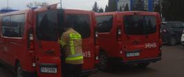 Na zdjęciu dwa pojazdy, czerwone straży pożarnej. Zachmurzenie duże. Przed pojazdami strażak w ubraniu specjalnym.