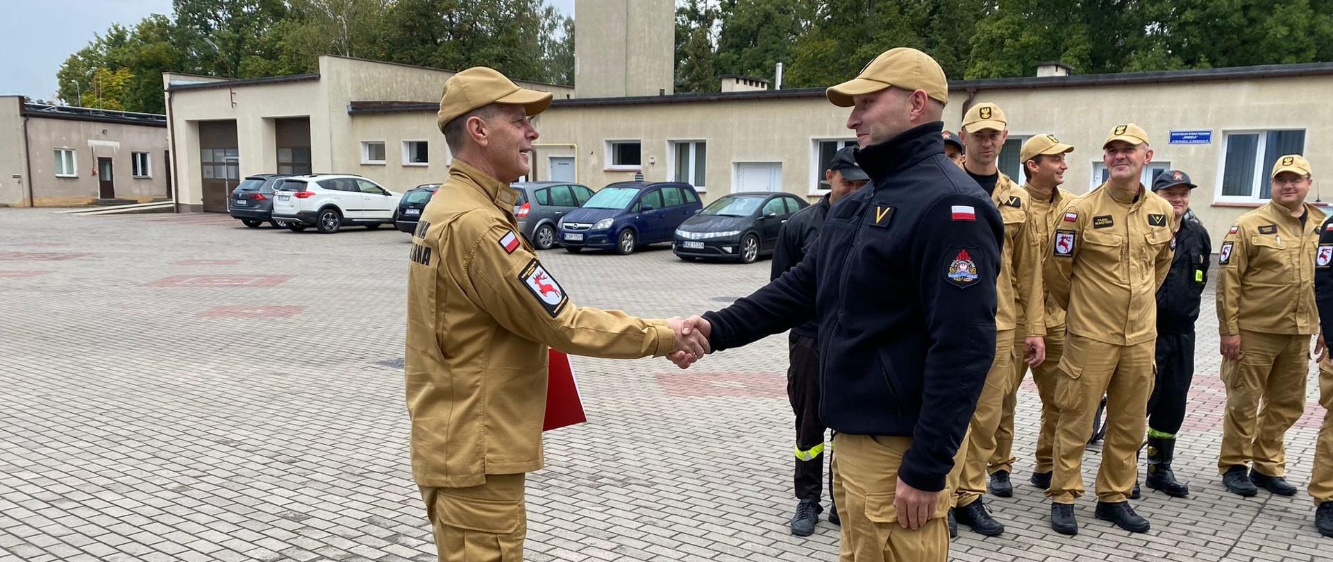 Zdjęcie przedstawia st. bryg. Andrzeja Góreckiego Komendanta Powiatowego PSP w Brodnicy wręczającego ogn. Mariuszowi Boruczkowskiemu list gratulacyjny wraz z informacją o przyznaniu nagrody z okazji jubileuszu pracy zawodowej i służby w szeregach Państwowej Straży Pożarnej. Za nimi po prawo strażacy podziału bojowego. Dalej samochody przed budynkami Komendy.