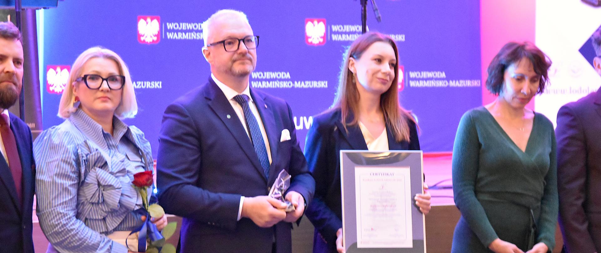 Wojewoda warmińsko-mazurski, dyrektor generalna oraz pracownicy Warmińsko-Mazurskiego Urzędu Wojewódzkiego pozują do wspólnego zdjęcia. Dyrektor generalna trzyma certyfikat, a wojewoda statuetkę. Wszyscy stoją na tle ścianki z oznaczeniem Wojewody Warmińsko-Mazurskiego.