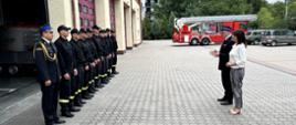 Z lewej pododdział strażaków stojący w rzędzie wzdłuż ściany budynku JRG2. Z prawej Pan Komendant i Pani Poseł przemawiający do strażaków.